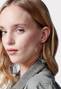 Pendientes de aro plateados con una gran bola decorativa, llevados por una persona con cabello rubio, frente a un fondo liso.