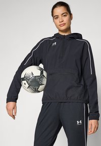 Svart hettejakke med hvite detaljer, halvmeter glidelås foran og elastisk liv, kombinert med svarte joggebukser, og holder en mønstrete fotball.