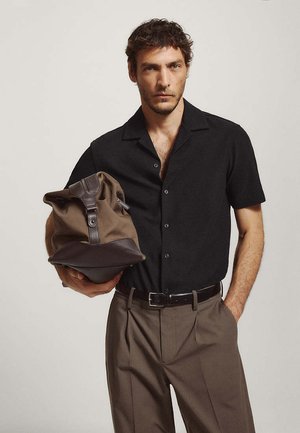 Homme portant une chemise à manches courtes noire et un pantalon marron tient un sac marron avec des accents en cuir avec son bras gauche sur un fond uni.
