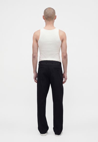 Homme à la tête rasée portant un débardeur blanc et un pantalon noir, debout de dos sur un fond blanc uni.