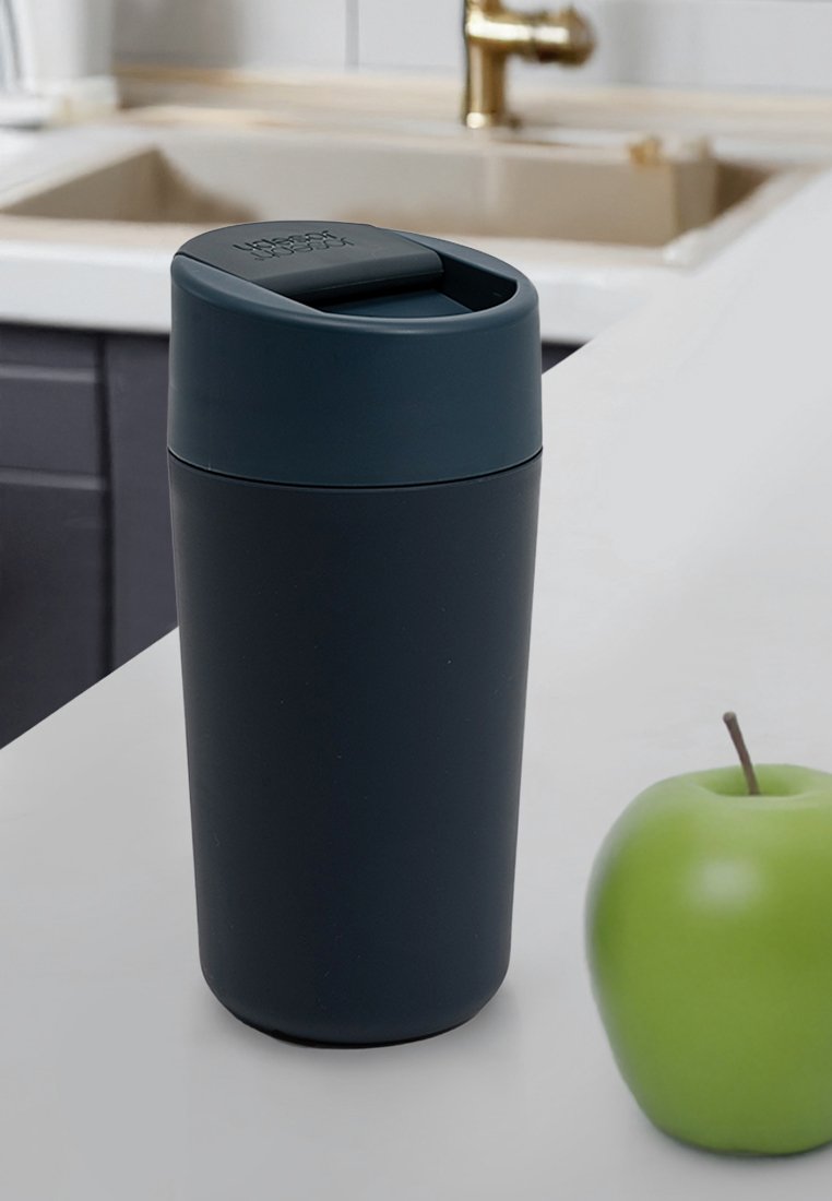 Granatowy, izolowany kubek podróżny o zakrzywionym kształcie, z płaską pokrywką i matowym wykończeniem. Obok niego na białym blacie leży zielone jabłko.
