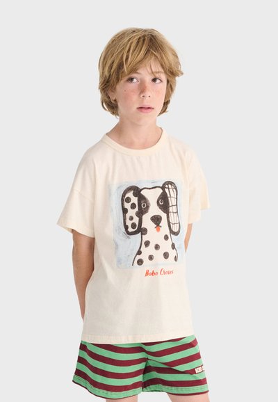 Bambino biondo con maglietta crema con illustrazione di un cane dalmata e pantaloncini a righe verdi e marroni, in piedi con le mani dietro la schiena.