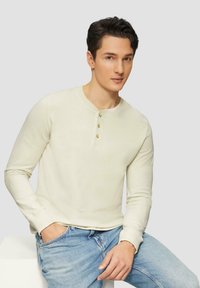Langärmliges cremefarbenes Henley-Shirt mit drei goldenen Knöpfen am Ausschnitt, glattem Textur und taillierten Ärmeln, kombiniert mit hellblauen Jeans.