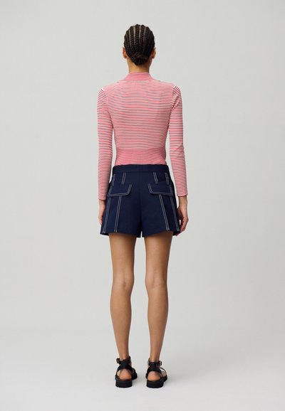 Maglietta a maniche lunghe a righe rossa e bianca, abbinata a pantaloni corti blu navy con tasche frontali e accenti di cucitura bianca. Sandali neri completano il look.
