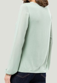 Top verde chiaro a maniche lunghe con una texture morbida, colletto rotondo e maniche semitrasparenti, abbinato a jeans scuri.