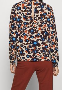 Halenka s dlouhým rukávem v béžové barvě s barevným leopardím potiskem, který obsahuje modré, oranžové a hnědé skvrny; pružné manžety a otvor na zádech.