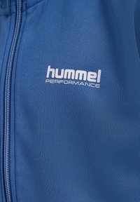 Niebieska kurtka sportowa z zamkiem błyskawicznym, wykonana z teksturowanego materiału, z białym logo "hummel PERFORMANCE" z przodu.