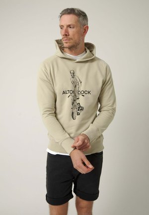 Hombre con cabello gris corto, viste una sudadera con capucha beige con un dibujo de motocicleta y pantalones cortos negros, ajustándose la manga contra un fondo liso.