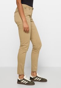 Personne portant un pantalon beige slim et des baskets noires et blanches, debout de côté sur un fond blanc uni.