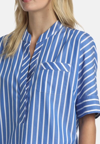 Femme portant une chemise rayée bleu et blanc verticalement, avec un col sans revers et une poche poitrine, montrant une partie de son cou et de sa bouche.