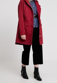 Parka in cotone rosso con cappuccio, dotata di tasche frontali e coulisse. Abbinata a una camicia di denim e pantaloni a vita alta in velluto nero. Stivaletti neri alla caviglia.