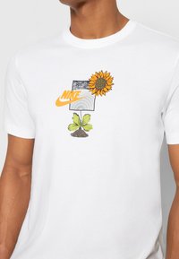 Camiseta blanca de algodón con un diseño gráfico que presenta un girasol amarillo, hojas verdes, un terreno de tierra y una forma abstracta en blanco y negro, además de un logo de Nike.