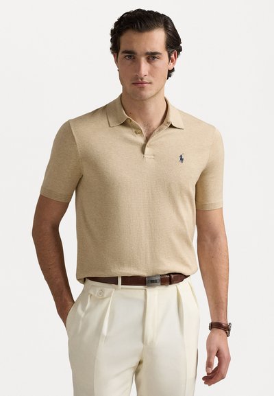 Polo shirt bej cu mâneci scurte și guler, având un mic logo bleumarin pe piept. Purtați cu pantaloni de culoare crem și o curea maro.