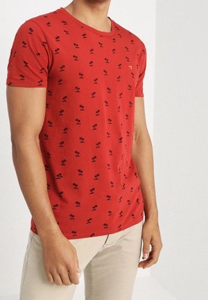 Jeune homme portant un t-shirt rouge à manches courtes avec de petits motifs de palmiers noirs, associé à un pantalon beige clair, debout contre un mur blanc.
