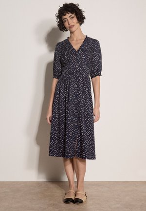 Donna con capelli corti e ricci indossa un vestito midi blu navy con pois chiari, maniche a sbuffo e scarpe basse beige e nere.