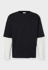 Μαύρο κοντομάνικο μπλουζάκι φορεμένο πάνω από λευκό πουκάμισο με μακριά μανίκια, απλωμένο σε λευκό φόντο.