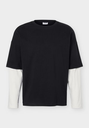 Maglietta nera a maniche corte sovrapposta a una camicia bianca a maniche lunghe, mostrata distesa su uno sfondo bianco.
