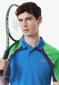 Blå och grön pikétröja med krage, i ett texturerat material, med kontrasterande marinblå detaljer och en liten logotyp. En tennisklubba hålls i handen.