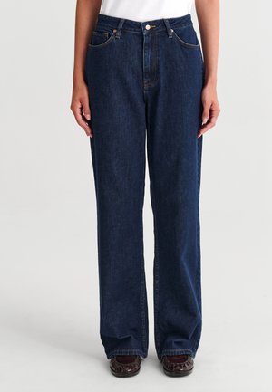 Personne portant un jean bleu foncé droit avec poches avant et un haut blanc, debout les mains détendues le long du corps.
