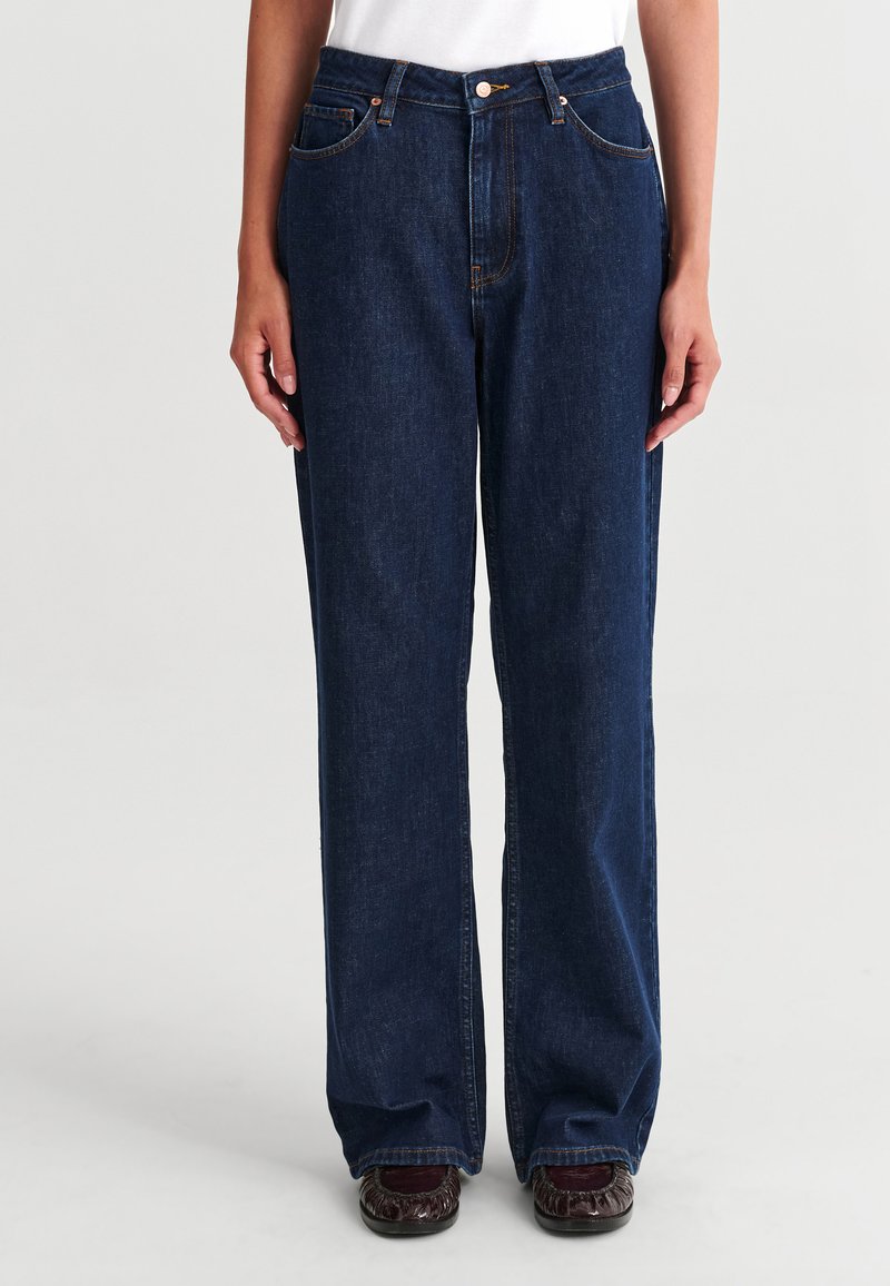 Personne portant un jean bleu foncé droit avec poches avant et un haut blanc, debout les mains détendues le long du corps.