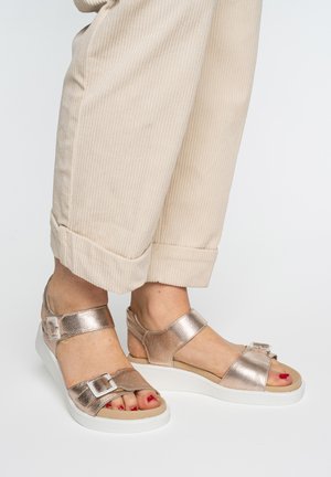 Feet wearing metallic rose gold sandals with white soles and buckles, paired with beige rolled-up corduroy pants.