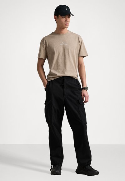 Μπεζ βαμβακερό t-shirt με γραφικό σχέδιο, συνδυασμένο με μαύρο cargo παντελόνι που διαθέτει πολλές τσέπες, φορεμένο με μαύρα αθλητικά παπούτσια και καπέλο.