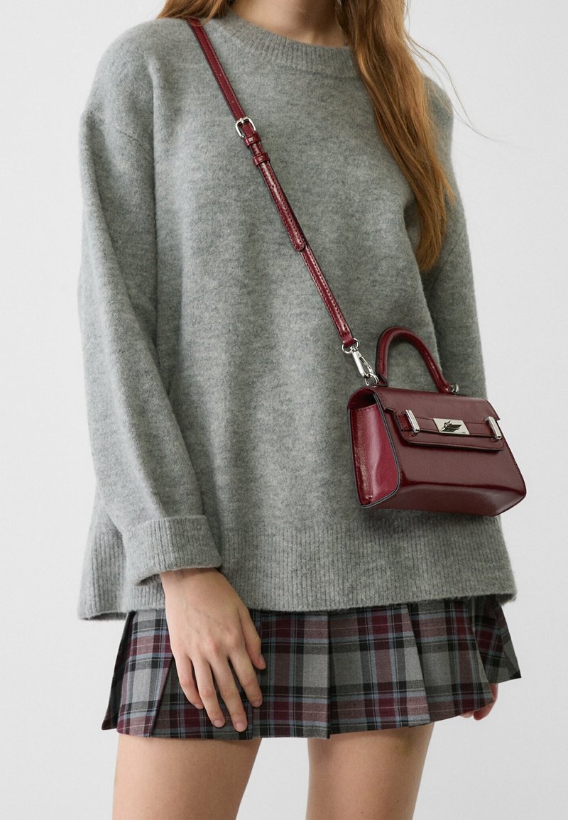 Personne portant un pull gris, une jupe à carreaux et un petit sac bandoulière en cuir bordeaux avec une poignée supérieure et une fermeture argentée.