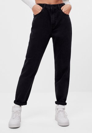 Person trägt schwarze Jeans mit hohem Bund und aufgerollten Bündchen sowie weiße High-Top-Sneaker, Hände in den Taschen, vor neutralem Hintergrund.