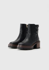 Black leather ankle boots with chunky brown platform heels, side zipper, and decorative back straps on neutral light background.