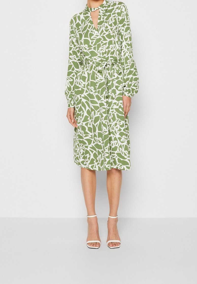 Femme portant une robe verte mi-longue avec des motifs de feuilles blanches, des manches longues, un col en trou de serrure et une ceinture à la taille, associée à des talons blancs.