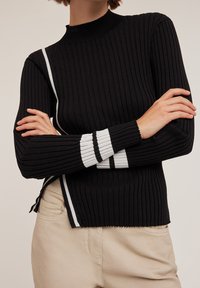 Czarny sweter prążkowany z wysokim dekoldem, z białymi paskami po bokach i wykończeniu rękawów, w połączeniu z jasnobeżowymi spodniami.