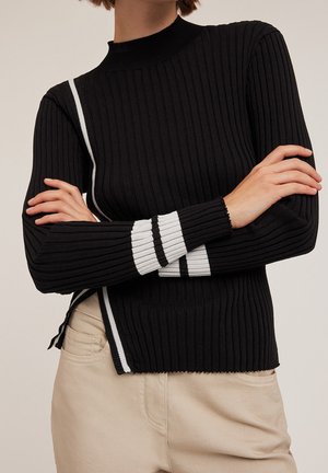 Pull noir côtelé avec un col montant, comportant des rayures blanches sur les côtés et les poignets, associé à un pantalon beige clair.