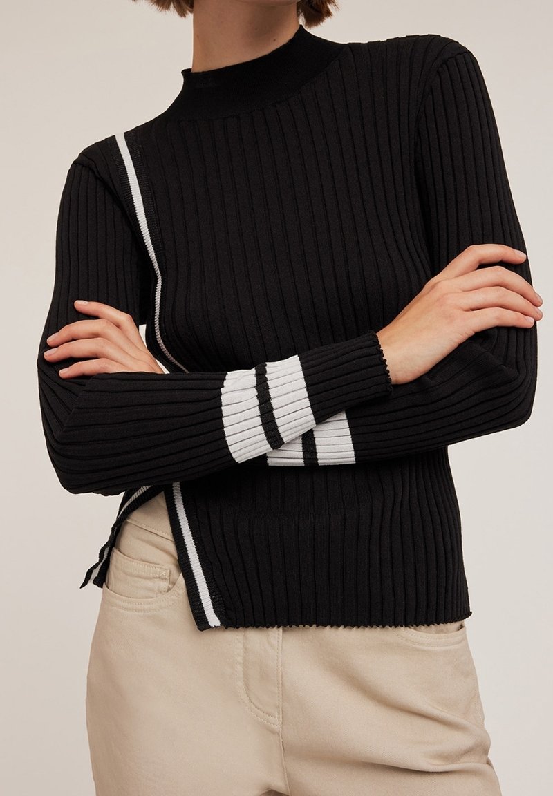 Czarny sweter prążkowany z wysokim dekoldem, z białymi paskami po bokach i wykończeniu rękawów, w połączeniu z jasnobeżowymi spodniami.