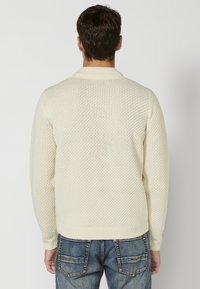 Crème gebreide trui met een textuurpatroon, ronde halslijn en geribbelde manchetten. Gecombineerd met blauwe denim jeans met achterzakken.