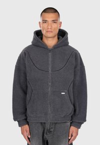 Uomo che indossa una felpa in pile grigio scuro con zip e tasche anteriori, pantaloni neri, in piedi contro uno sfondo chiaro e semplice.