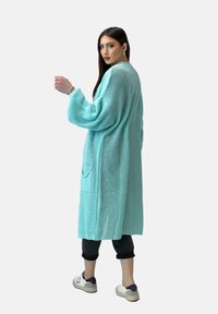 Cardigan lungo aperto in maglia turchese chiaro con tessuto testurizzato, due tasche laterali e maniche morbide, abbinato a pantaloni neri e sneakers bianche.