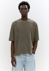 Oliven grøn ribbet strik T-shirt med korte �ærmer og rund halsudskæring, parret med lyseblå jeans med hvid broderet tekstdetalje.