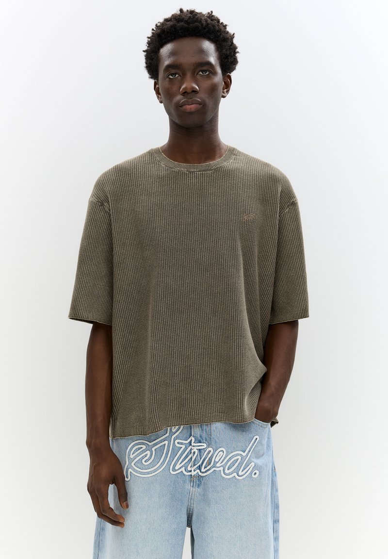 Olijfgroene geribbelde trui met korte mouwen en een ronde hals, gecombineerd met lichtblauwe jeans met witte geborduurde tekstdetails.