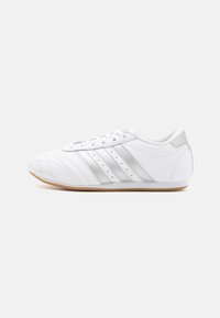 White leather athletic shoe with a rounded toe, silver stripes, and perforated detailing on the sides. Tan rubber sole. Standard lace-up design.