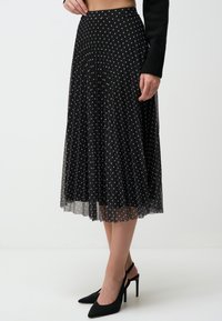 Gonna nera plissettata con pois bianchi, realizzata in materiale trasparente, dotata di una vita aderente e una silhouette fluida.