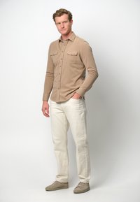 Camicia marrone chiaro a maniche lunghe con due tasche sul petto, abbinata a jeans avorio e scarpe da ginnastica beige, su uno sfondo semplice.