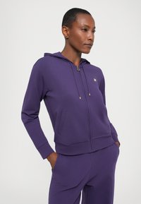 Paarse zip-up hoodie gemaakt van zacht materiaal, met een capuchon, voorzakken en gouden hardware. Gladde textuur en aansluitende pasvorm.