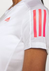 Vit sporttröja med rosa Adidas-logotyp och tre vertikala ränder. Materialet har en texturerad, andningsbar meshtyp.