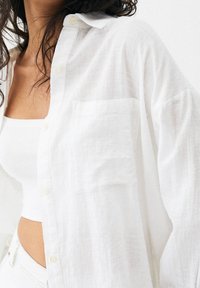 White linen shirt with a relaxed fit, featuring a collar, front pocket, and button details. Worn over a white fitted tank top.