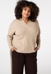 Beige gebreide trui met een V-hals en lange mouwen, gecombineerd met donkerbruine broek met zijstrepen. Zachte textuur en losse pasvorm.