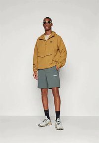 Khakifarbener Pullover mit einer Vorderasche, kombiniert mit grauen Shorts. Das Model trägt eine Sonnenbrille und helle Sneaker und steht vor einem schlichten Hintergrund.