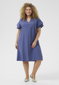 Kleid mit kurzen Ärmeln in knielanger Länge aus einfarbigem Lavendelstoff, mit einem V-Ausschnitt und geschichteten Ärmeln. Kombiniert mit hellen flachen Schuhen.