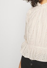 Bílá bavlněná blůza s detaily ve stylu eyelet, volánkovým límcem, nařaseným pasem a balonovými rukávy. Texturována malými perforacemi a pruhy.