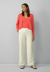 Korallenfarbener gestrickter Cardigan über einem passenden Top, kombiniert mit weiten beigefarbenen Hosen im Karomuster. Das Outfit zeichnet sich durch eine strukturierte Oberfläche und minimalistische Details aus.