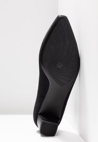 Chaussure noire à bout pointu avec dessus en tissu texturé, semelle inclinée montrant le motif de la bande de roulement, et talon carré bas sur fond blanc.